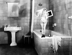 Thelma Todd stands on the edge of a large bathtub in an oversized