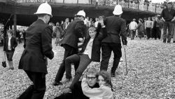 the60sbazaar:  A mod couple on the beach as a riot goes on around