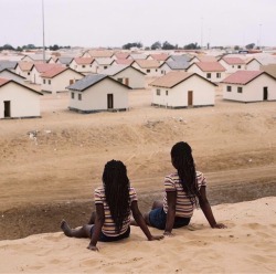 forafricans:Two girls in matching clothes sit on a hill of sand.