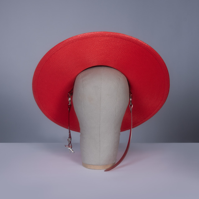 Wide brim hat in red faux straw with translucent red pvc buckle hat band and harness strap details, displayed on a mannequin head against a gray background.