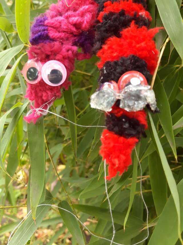 A pink string worm named Roxy and a red and black string worm named Dave sitting side by side on a green bamboo bush