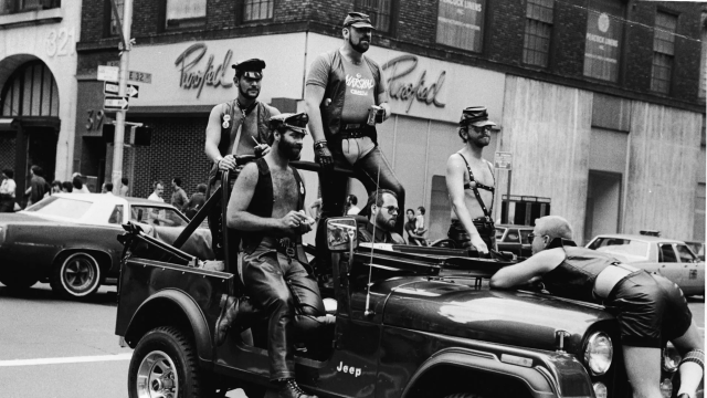 Black and white photo of bare-chested leather daddies riding in a vehicle during a parade