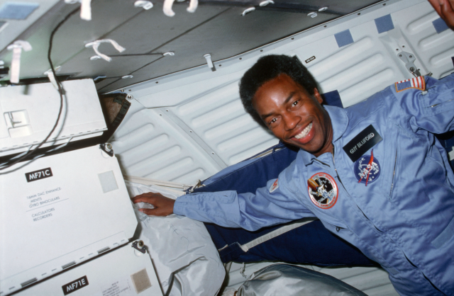 Guy Bluford, an African American man, floats near storage inside the Challenger spacecraft. He has one hand on a shiny gray bag with markings on it, and the other is nearly off-screen on the right. He wears a powder blue jumpsuit that has various zippers on it, as well as NASA, mission, and flag patches. He is looking directly at the camera while smiling. Credit: NASA