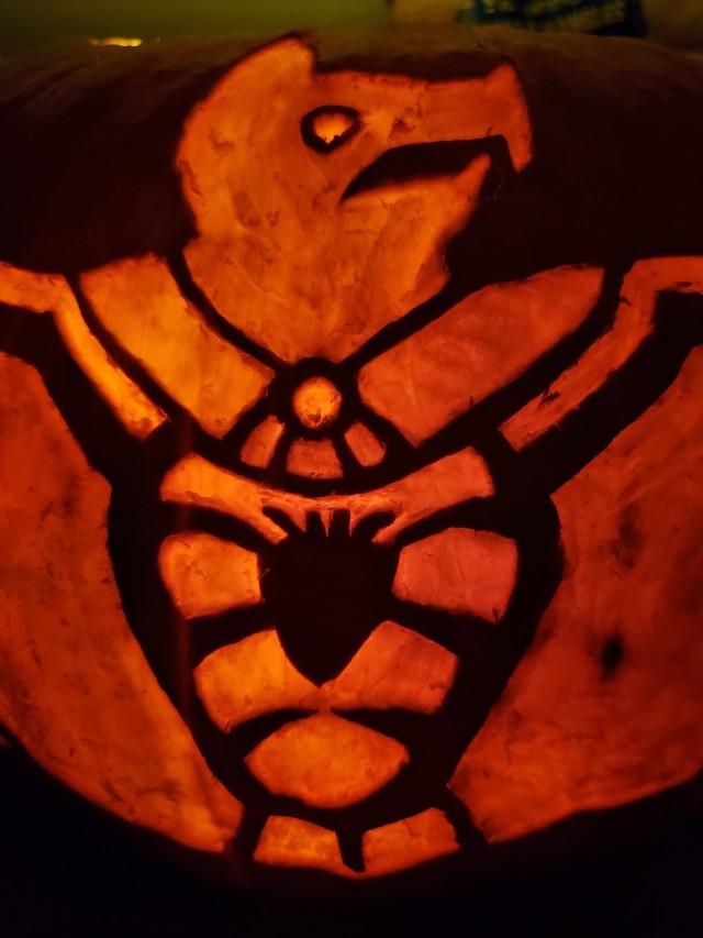 Up close front view of a pumpkin carved so that its flesh glows bright orange without being all the way cut through. The image is of a Mesoamerican styled image of a bat, with an open rib cage showing the mexica styled heart. The circular eye is slightly brighter than the rest of its body.