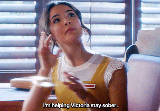 GIF FROM EPISODE 1X07 OF NANCY DREW. BESS SITS IN A BOOTH AT THE CLAW. SHE'S LOOKING UP, TALKING TO SOMEONE STANDING OUT OF FRAME. SHE SAYS "I'M HELPING VICTORIA STAY SOBER" AND POINTS OFF-CAMERA TO WHERE VICTORIA IS SITTING.