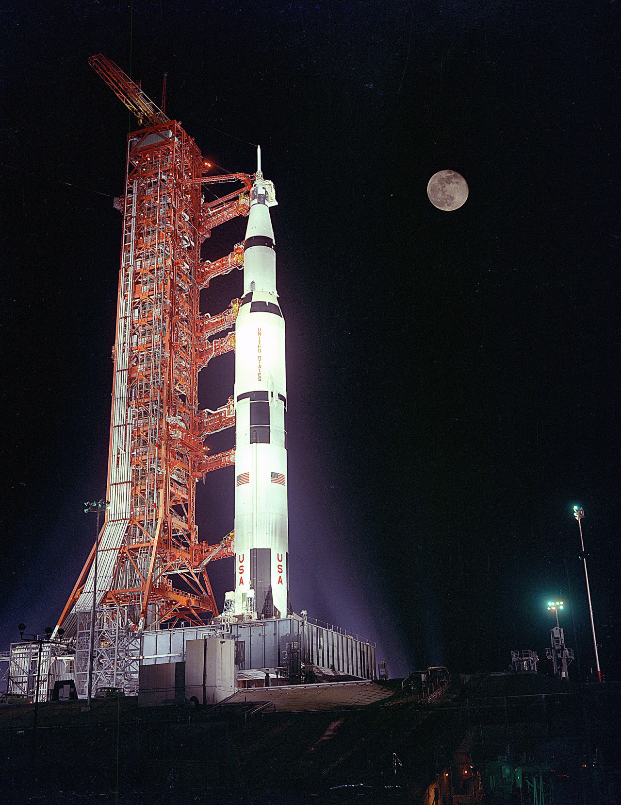 The Apollo 17 Space Vehicle sits poised beneath a full moon on Launch Pad 39A at the Kennedy Space Center during the launch countdown. The Saturn V rocket is mostly white, with several black patches, American flags, and the letters “USA” on its side. It is connected to an orange launch tower on the left. Credit: NASA