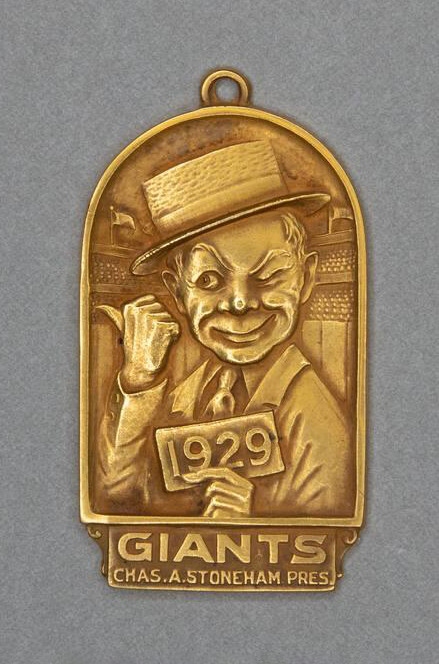 A 1929 New York Giants baseball season admission pass in the form of a small, arch-shaped gold medal with a loop at the top. On the obverse side of the pass is a caricature image of the head and shoulders of a winking man wearing a suit and a straw hat. In his left hand he holds a card reading "1929" and his right thumb points off to the side of the pass. A baseball stadium is seen behind him. Text at the bottom of the pass reads: GIANTS / CHAS. A. STONEHAM PRES.