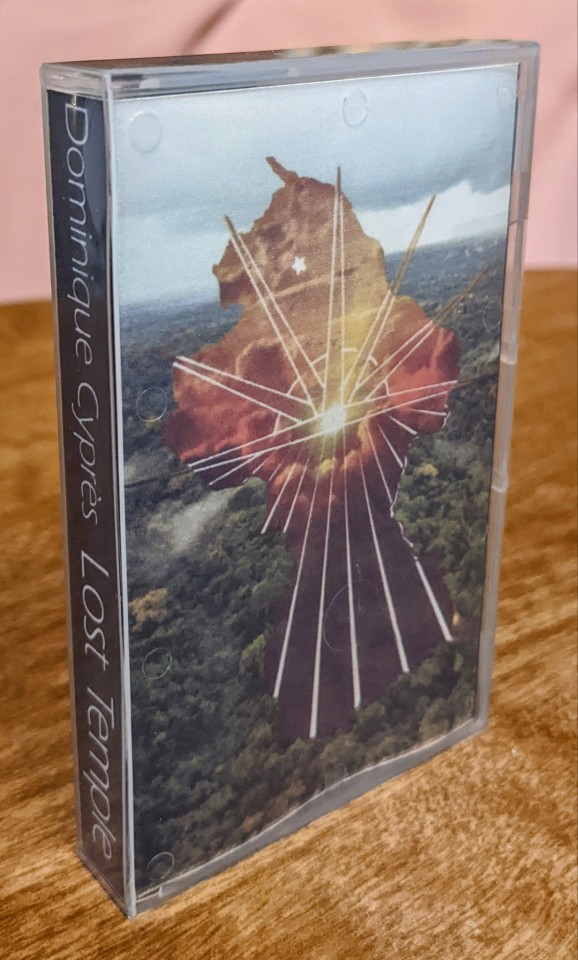 Front and spine of the cassette box. The front shows the Jonestown logo, a sunburst design on an outline of Guyana, with a small star indicating the location of Jonestown. Behind the logo is an aerial photo of misty Guyanese jungle. The spine bears the name Dominique Cyprès and the album title Lost Temple.