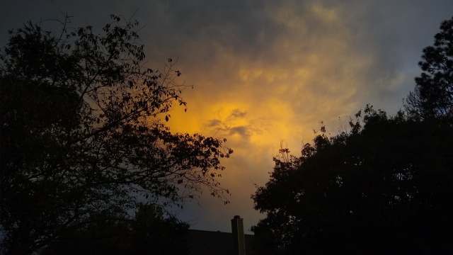 A photo of a sunset, focused on a yellow cloud with the chimney of a house and trees also visible