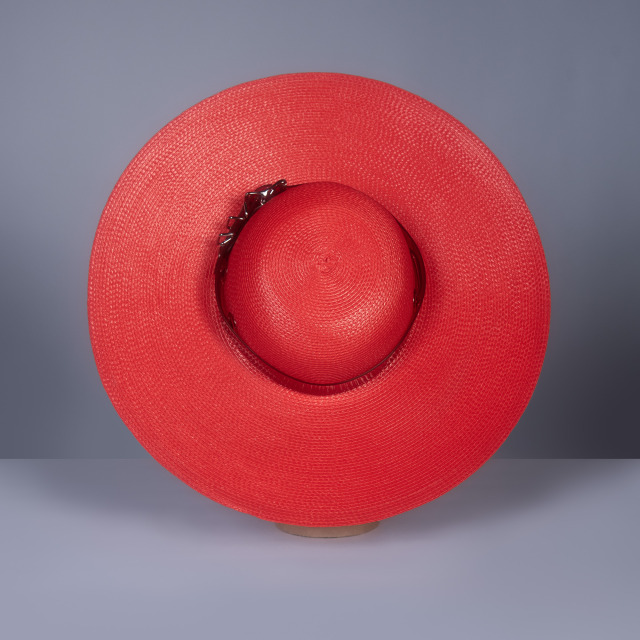 Wide brim hat in red faux straw with translucent red pvc buckle hat band and harness strap details, displayed on a mannequin head against a gray background.