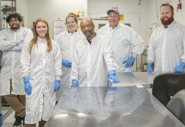 Two Black men, two white women, and two white men each stand in white lab coats and blue gloves. All are smiling. They are in a small room with silver metallic tables, one of which in the foreground reflects some of their likenesses. Credit: NASA/Chris Gunn 