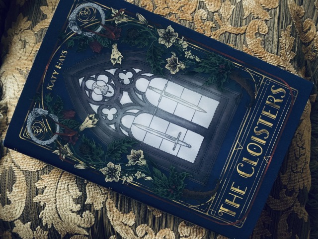 Pictured is the Fox & Wit special edition of THE CLOISTERS, with cover art depicting a stained glass windows, flowers, and ouroboros symbols, shown against a gold fabric background. Photo by AHS.