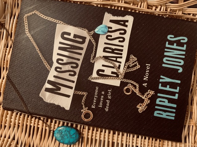 Pictured is MISSING CLARISSA (with cover art depicting a necklace with a turquoise charm) against a wicker backdrop, beside a necklace with a turquoise charm. Photo by AHS.