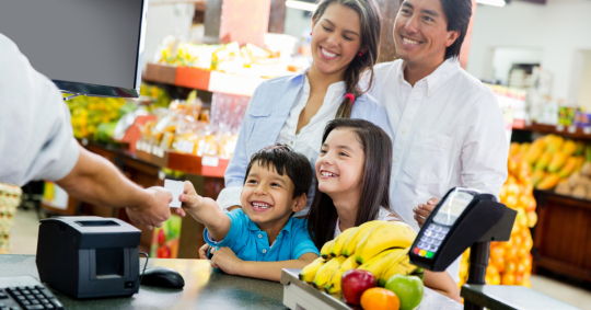 Family smiling at grocery store check out. Little boy cheerily hands credit card to cash register clerk.