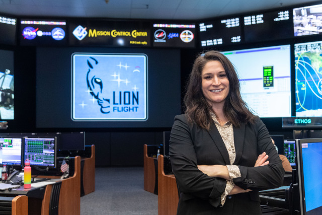 Chloe Mehring, a woman with shoulder-length brown hair, poses for a picture in the Mission Control Center at NASA’s Johnson Space Center in Houston. She wears a black blazer, and her arms are crossed as she smiles. Behind her are several desks lining an aisle. On the desks are many computer screens. Large screens line the walls with the logos of NASA and other space agencies, times, maps, and more information. Credit: NASA