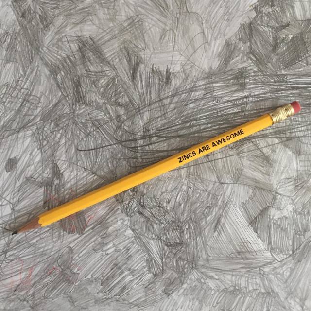 Image of a yellow pencil with the words ZINES ARE AWESOME embossed in black. The pencil sits on a grey background of random pencil scrawls. 