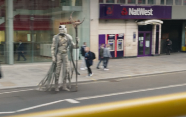 Creepy bird mummy with a crescent moon staff standing on the pavement as people pass by. No one else can see him except the protagonist. He's standing outside a Natwest bank which you can see in the background