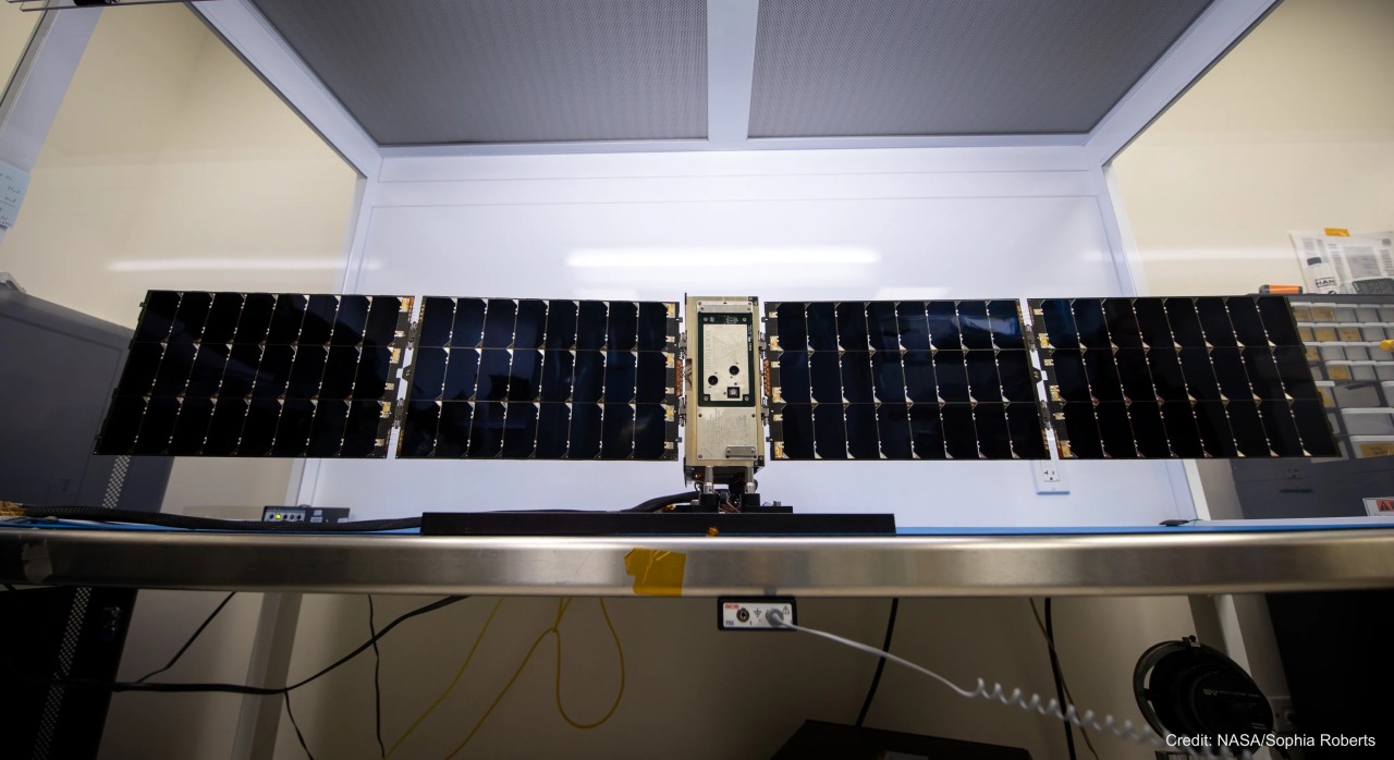 This image shows a small spacecraft on a table enclosed on all sides except the one facing the camera. The sides of the enclosure are clear while the top has two dark gray panels with a light gray frame. The backside is also gray and reflects a strip of light from the room. The spacecraft’s body is a vertical golden rectangle. Shiny black solar panels extend to either side and are much wider than the spacecraft itself. There are a few wires connected to the table, which are visible underneath it. It’s watermarked, “Credit: NASA/Sophia Roberts.”