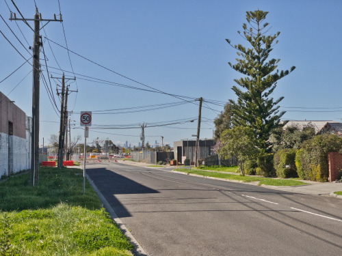 The view down derby road facing east in August 2021. A typical suburban street with houses and a smattering of trees on the right and a factory wall on the left. There are a smattering of orange construction Barries in the middle ground. There is an intersecting street, Thorpe Street. This corner has a real estate sales office on it. The CBD skyline can just be seen in the background. The sky is blue and clear.