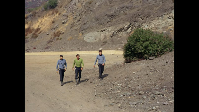 ID: A screenshot from Star Trek TOS, of Spock, Kirk, and Bones walking on a dirt covered floor. Behind them is dirt-covered hills populated with dead grass and to the right is a green bush. End ID]