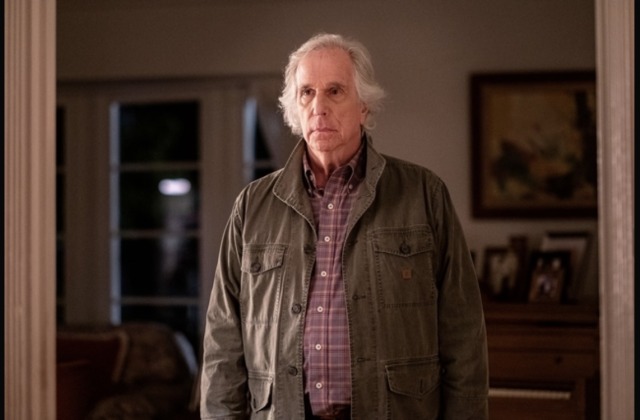 Henry Winkler as Gene Cousineau in a plaid shirt and olive jacket. He is standing in a door/archway with artwork, windows and furniture behind him