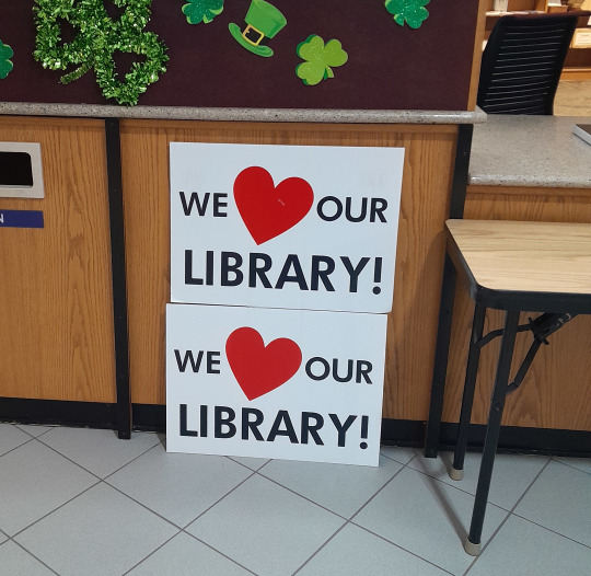 A white yard sign that reads We heart our library. The heart is red