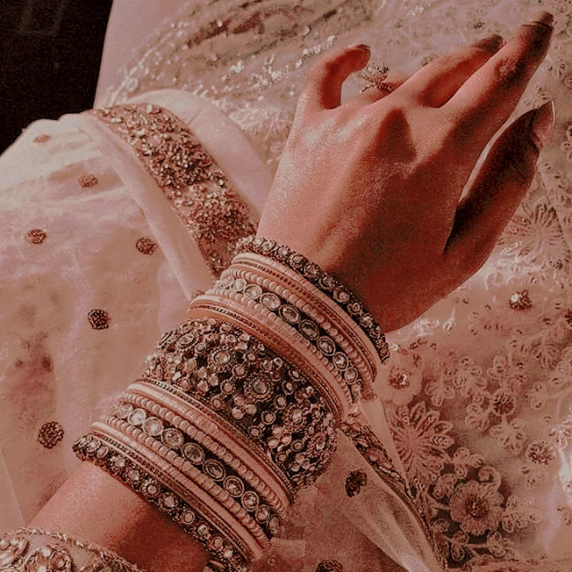 the arm of an indian woman, adorned with multiple pink and russet gold bracelets, pictured against her elaborate pale pink and gold embroidered lehenga.