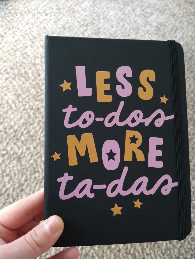 a black notebook with the words "LESS to-dos MORE ta-das" in purple and orange, surrounded by stars