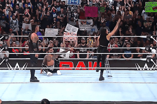 A gif of Rhea Ripley. She is spinning on one leg holding a crutch and lifting her arm up and down to "yeet" to Jey Uso's theme music. Jey Uso and Damian Priest are also seen in the frame.