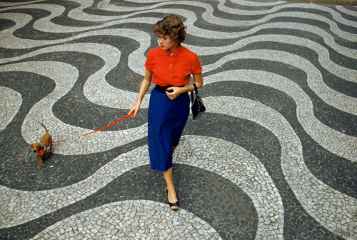 A woman walks a dachshund across pavement with undulating wave patterns in Rio de Janeiro, Brazil, M
