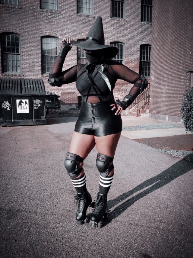 black person wearing a black witch hat, face mask, black sheet top, black shiny shorts, knee pads, stripped black and white socks, roller skates tipping their witch hat and posing
