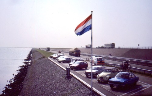 Parkeerplaats met Nederlandse vlag, verkeer en een 2CV, Afsluitdijk, Friesland, Nederland, 1984.