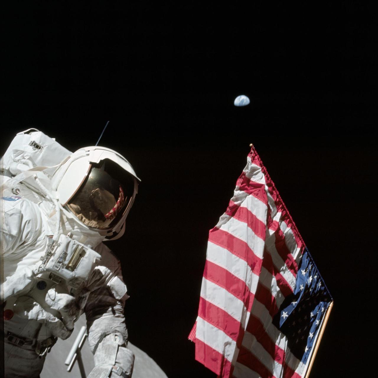 Astronaut Harrison Schmitt poses in a bulky white spacesuit on the Lunar surface next to an American flag. The Earth hangs in the black sky in the background, and fellow astronaut Eugene Cernan is seen in the reflection of Schmitt's golden visor. Credit: NASA