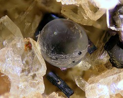 underthescopemin:  Fluorite, Sanidine, Augite Perfect round, transparent sphere of fluorite (a single crystal) with no crystal faces in a vug with sanidine and augite. Photo Copyright © Volker Betz 