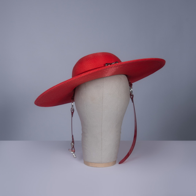 Wide brim hat in red faux straw with translucent red pvc buckle hat band and harness strap details, displayed on a mannequin head against a gray background.