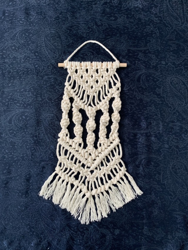 Photograph of a macrame wall hanging made from white cord on a wooden dowel, on a navy blue background. At the top is a triangle of square knots, followed by a v shape of diagonal clove hitch knots. Under this are five strands of half square knots which create a helix shape, followed by a v shaped row of forward and backward diagonal clove hitch knots. Then there is a v shape of alternating square knots and a last v shaped row of diagonal clove hitch knots. Under this, the cord is unravelled to give a frayed effect and cut in a v shape.