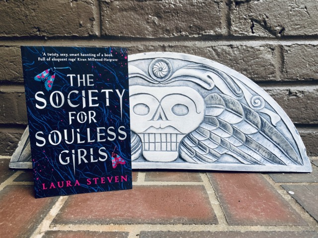Shown is THE SOCIETY FOR SOULLESS GIRLS (with cover art depicting colorful moths) sitting on bricks, against a brick background, next to a mock headstone with a skull and wings. Photo by AHS.