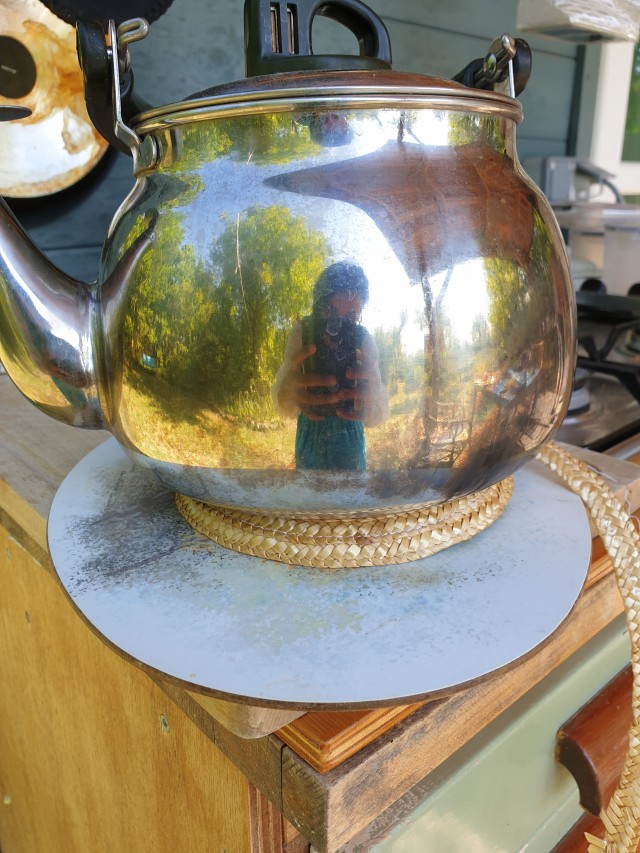 a shiny old school kettle on top of a straw round that will be the flat crowb of the hat, talia's distorted reflection is just visible in the kettle 