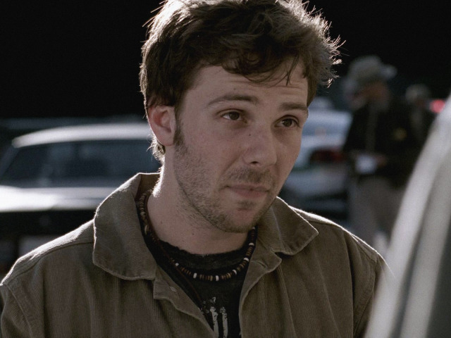 A shot from the chest up of Andy Gallagher looking forward. He is wearing a brown coat over a black shirt and a beaded necklace.