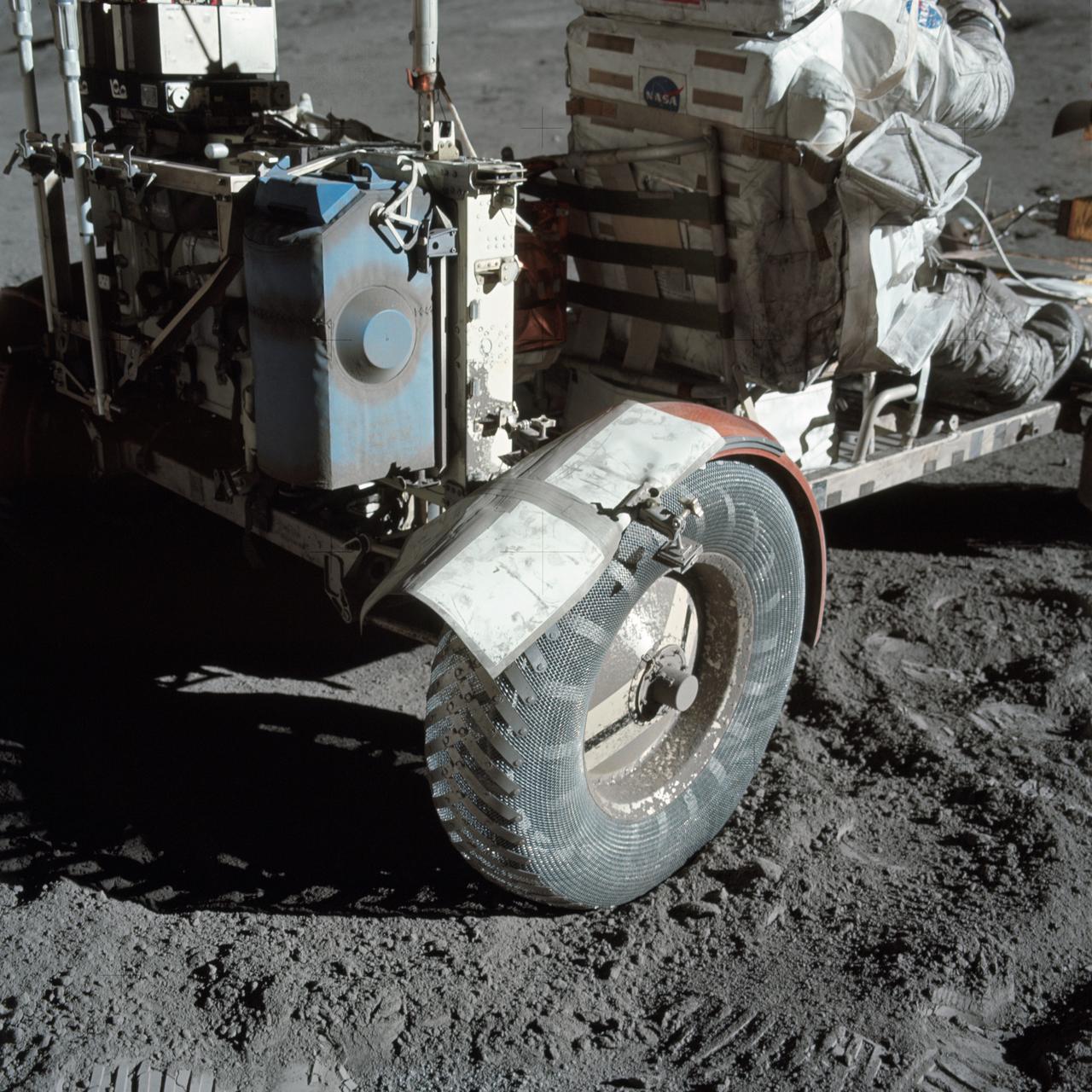 A close-up view of the rear right wheel of the Lunar Roving Vehicle (LRV) at the Taurus-Littrow. Note the makeshift repair arrangement on the fender of the LRV; a folded map is held in place parallel to the wheel with several strips of gray duct tape. Below the wheel, sunlight casts stark shadows on the dusty lunar surface. Credit: NASA