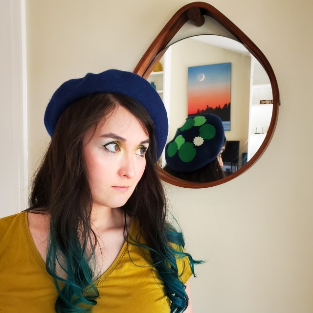 A photo of a woman wearing a golden shirt and a blue beret with a graphic of waterlilies on top. There is a mirror behind her.