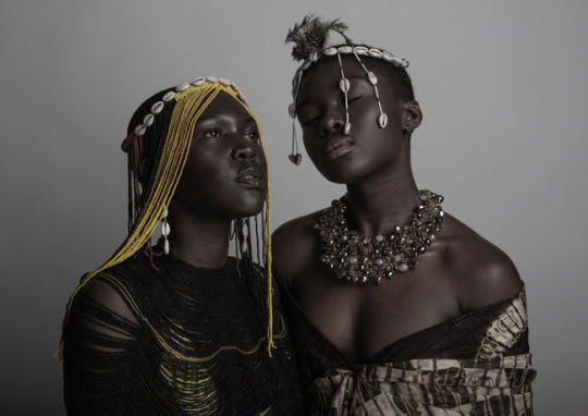 Image: Two women of African decent are pictured here. The woman on the left is looking up to her right wearing a black and red beaded blouse and a yellow and black beaded crown with cowrie shells on the top of the crown. On the right the women wears an over-the-shoulder draped top with a heavy beaded necklace and a cowrie shell beaded veil crown with black and white feathers. Her eyes are closed.