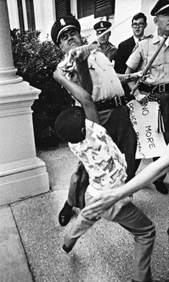 dichotomized:  In 1965, at Jackson, Mississippi, Matt Herron took an iconic and ironic image from the civil rights era as a white policeman rips an American flag away from a young black boy, having already confiscated his ‘No More Police Brutality’
