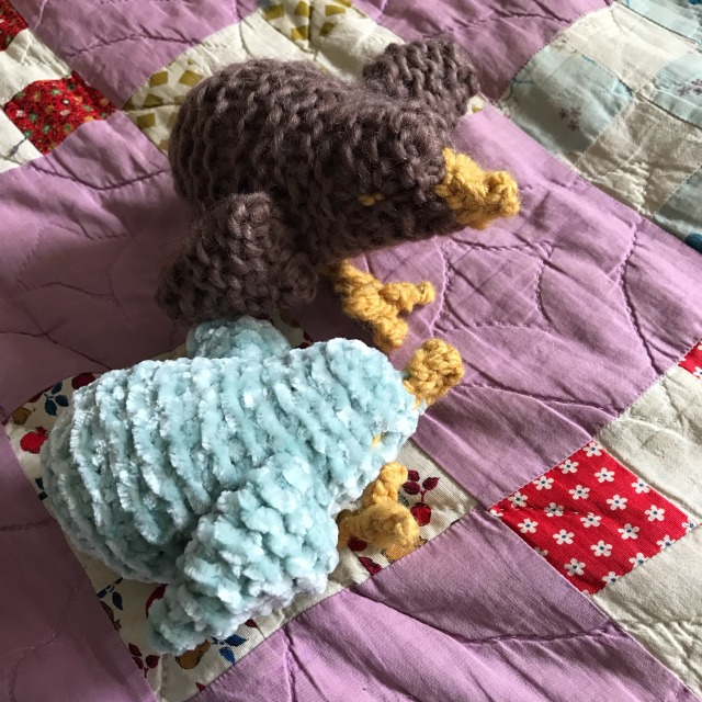 Two knitted birds with yellow beaks and feet sit on a purple quilt. One bird is brown, the other green.