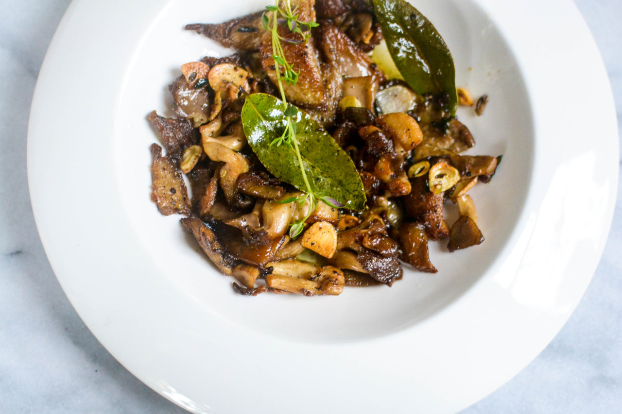 Overhead picture of sauteed oyster mushrooms with thyme, garlic, and bay leaves in a shallow white bowl on a marble slab