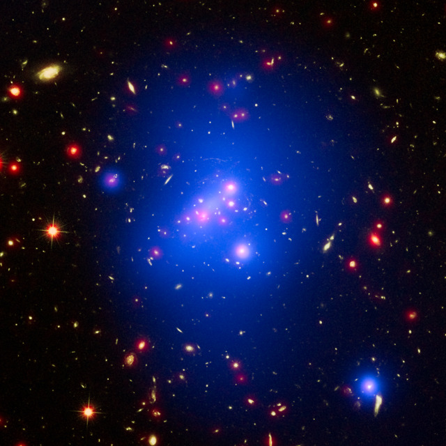 Vibrantly hued shapes speckle an image with a black background. Orbs glowing red, yellow, and blue are strewn across the frame, and a large, translucent blue haze dominates most of the center. Credit: NASA, ESA, and M. Brodwin (University of Missouri)