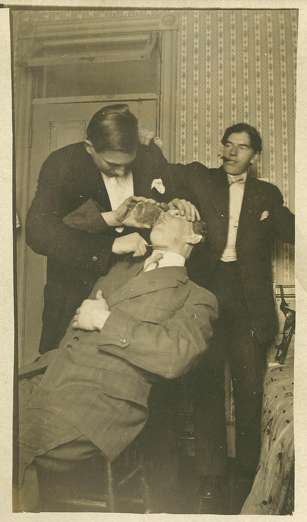 weirdvintage:  A man drinks while another man holds a straight razor to his throat,
