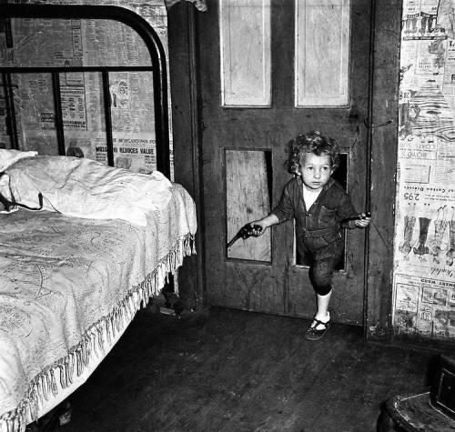 lettersfrombeachhead:mosertone:Coal  miner’s child using a hole in the door to enter a bedroom with a  smoking pipe in one hand and a gun in the other in Bertha Hill, West  Virginia. Photo by Marion Post Wolcott. 1938