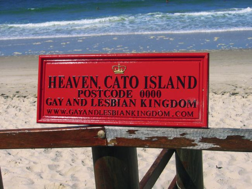 A red metal sign with a crown insignia at a beach reading "Heaven. Cato Island. Postcode 0000. Gay and Lesbian Kingdom."