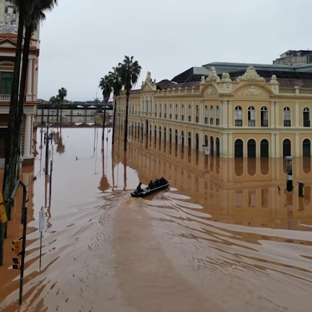 Picture taken in Porto Alegre. A historical building, the Public Market, is partially submerged in muddy water. A boat with four people is making a turn to the right to see the damage.